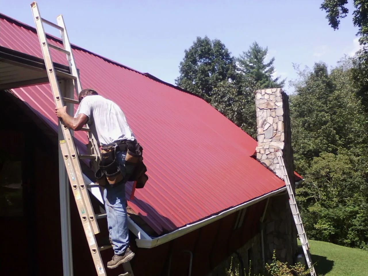 Roofer on a ladder beside a red metal roof and stone chimney — Soffit & Fascia Repair by Roofing Brothers in Minster